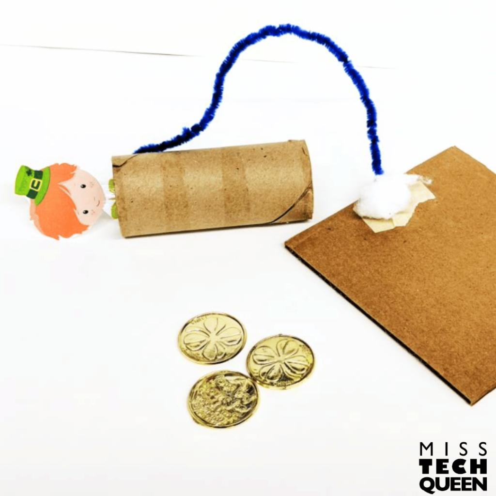 This image shows an example of leprechaun trap. A leprechaun figure is trapped in a toilet paper roll that is attached to a piece of cardboard by a pipe cleaner.